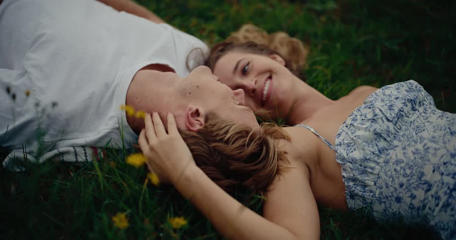 This video shows a romantic moment between a couple lying on their backs in the grass surrounded by small yellow flowers the woman with curly hair and wearing a light blue and white patterned dress is smiling and gazing lovingly at the man who is we