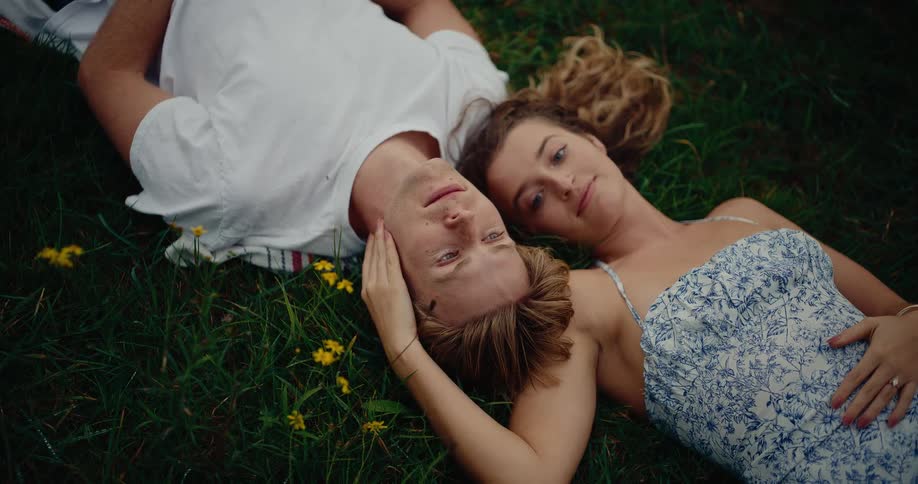 A young couple lies together in the grass looking into each others eyes with affection as the man rests his head on the womans shoulder while she gently caresses his face