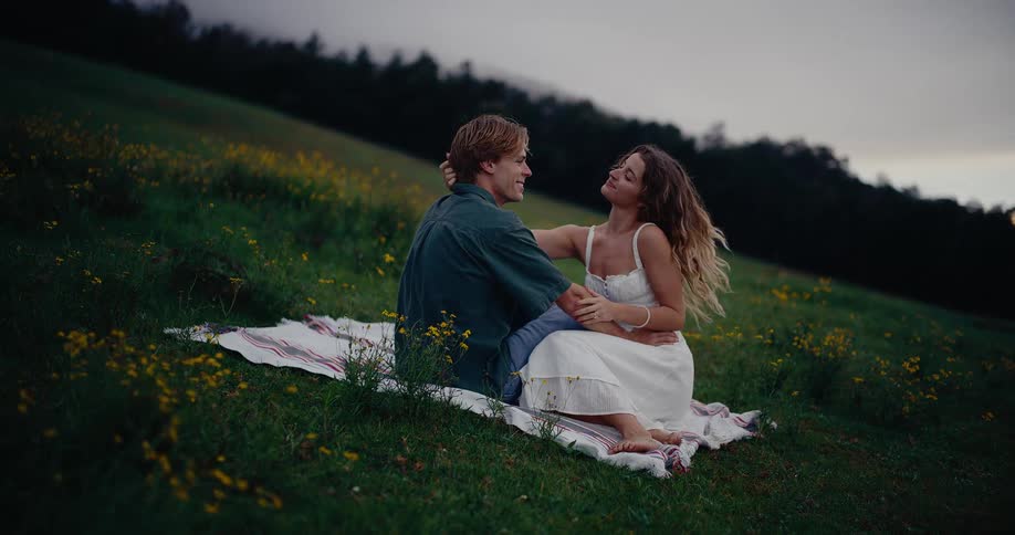 Woman touches her mans head while they sit on a blanket in a field near wildflowers on a summer day