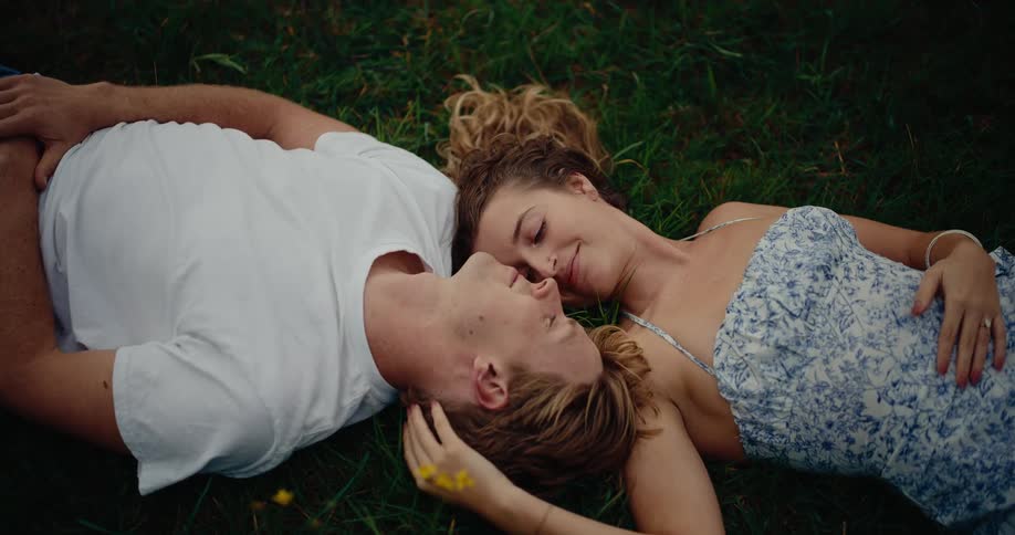 A couple lies together in the grass surrounded by dark green blades and small yellow flowers with the man gazing upward while the woman rests her head on his shoulder both dressed in light summer clothes
