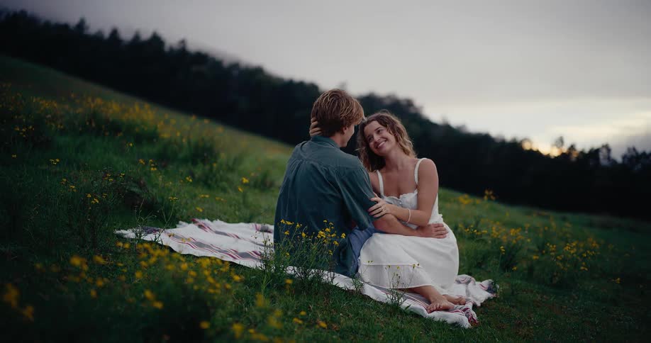 Woman smiles and leans her head against a mans while they sit on a blanket on a sloping field as a cross stands in the distance on a summer day