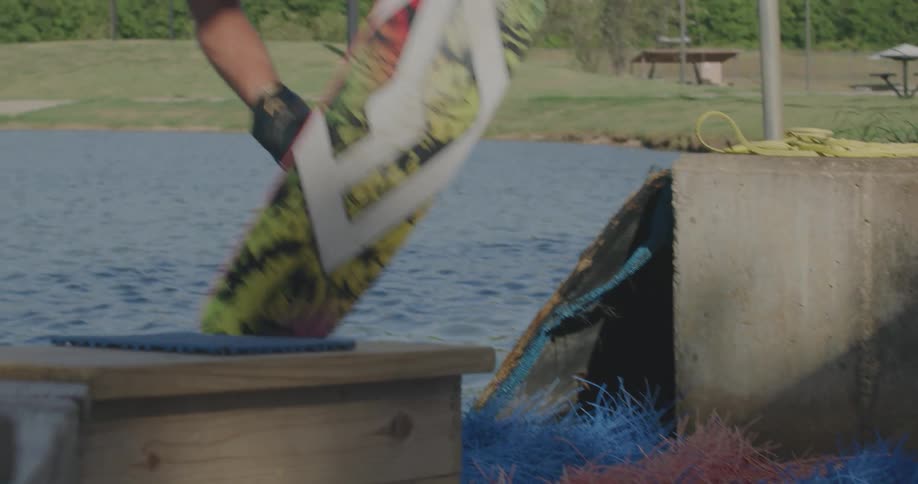 Standing on a dock lined with colorful matting a wakeboarder grips the tow handle and leaps into the lake instantly pulled forward across the rippling water by the overhead cable system