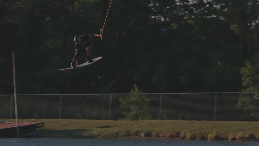 Man riding a wakeboard flies over a ramp and twists in midair as he holds a rope