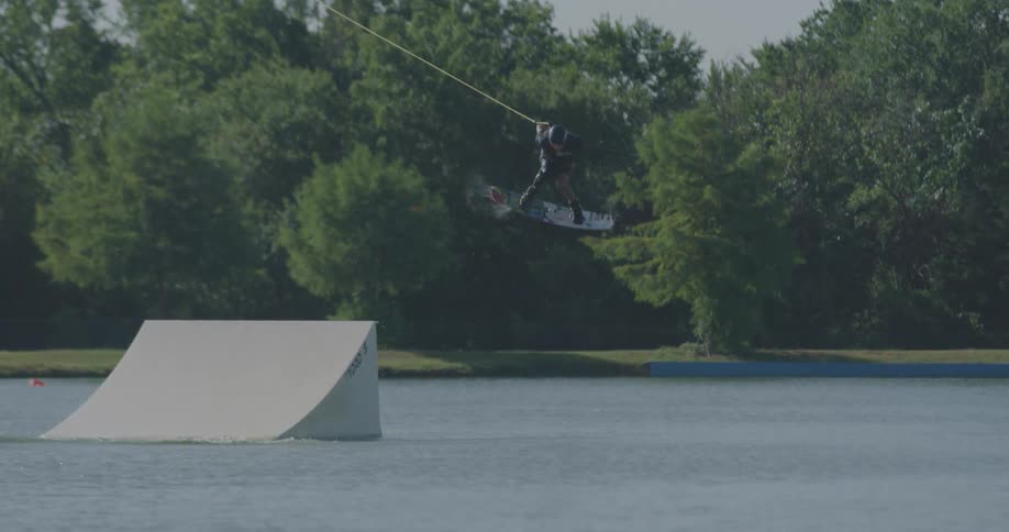 A wakeboarder successfully executes an aerial jump off a large white ramp at a cable park achieving significant air time before landing and creating a large spray of water