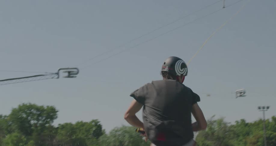 A wakeboarder performs a mid air trick over a lake with trees and grass in the background