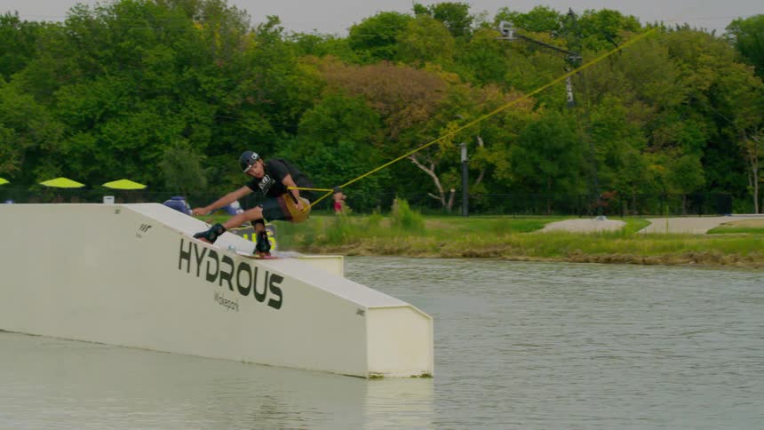 A wakeboarder crouched low with intense focus slides sideways along a white floating ramp labeled hydrous while gripping a taut yellow tow rope sending small ripples across the calm water