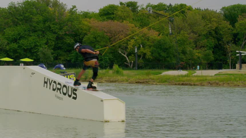A wakeboarder grinds along a cable park obstacle over a river with trees in the background