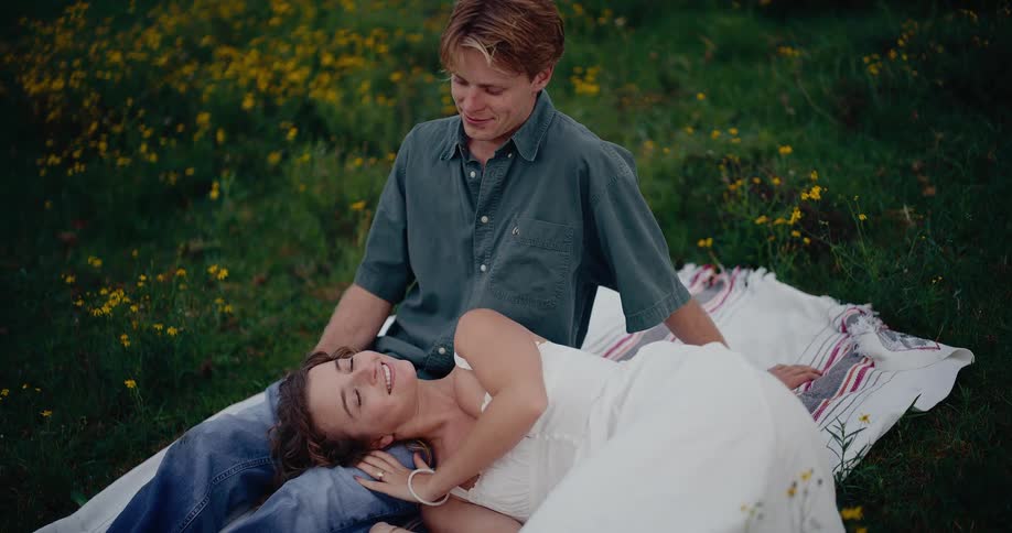 A man and woman smile at each other while sitting on a blanket in a meadow