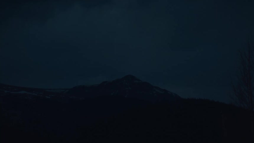 A dark rugged mountain silhouette against a deep blue overcast night sky creating a somber and isolated nighttime landscape