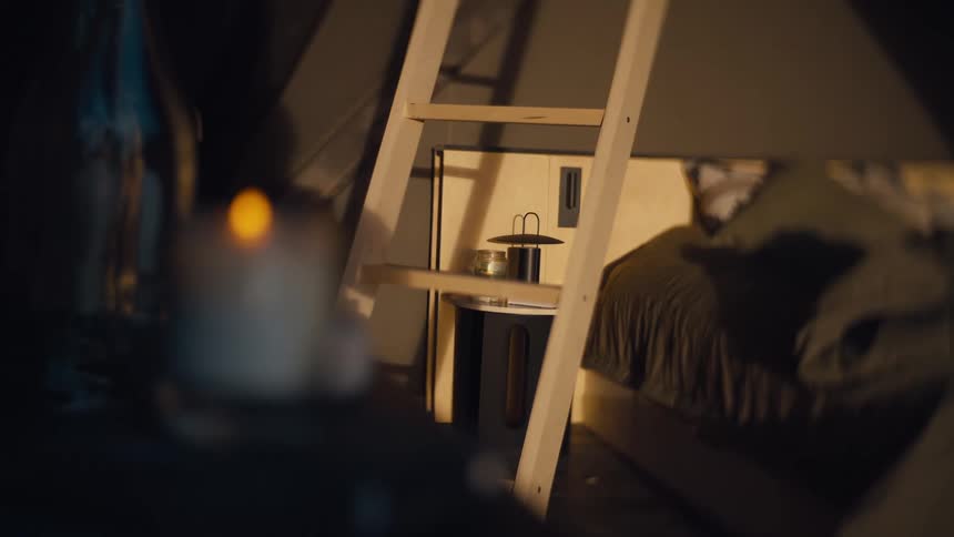 A warmly lit shallowfocus interior shows a wooden ladder leading up beside a neatly made bed with a small round table holding a lantern and objects in the background while a softly glowing candle