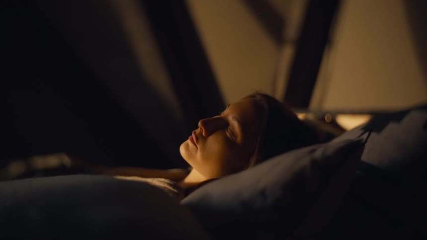 A young woman sleeps peacefully on a pillow in a dark room with a warm light source illuminating her face from the top right