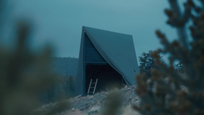 A wooden a frame tent stands alone on a rocky hillside at dusk surrounded by dark pine trees and brush