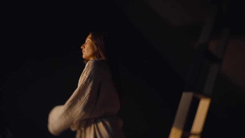 A woman walks through a dimly lit interior under warm soft lighting