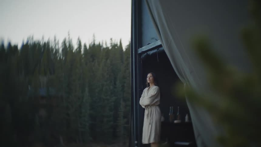 A woman wearing a light colored robe stands in the opening of a tent or structure looking out at a dense evergreen forest on a misty day