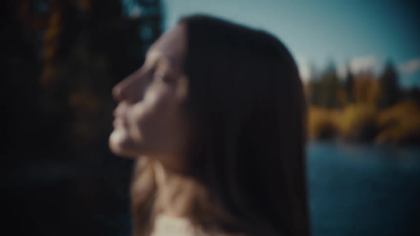A close view of a womans serene face eyes closed standing in soft sunlight outdoors with an out of focus autumn forest and river in the background