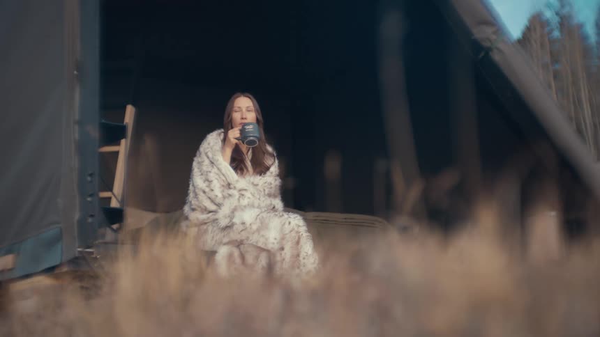 A woman wrapped in a white and gray blanket sits on the edge of a tent sipping from a black mug with a forest backdrop