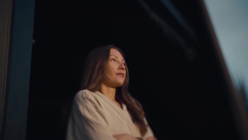 A young woman with long brown hair walks out of a building with her hands folded as the setting sun illuminates her skin