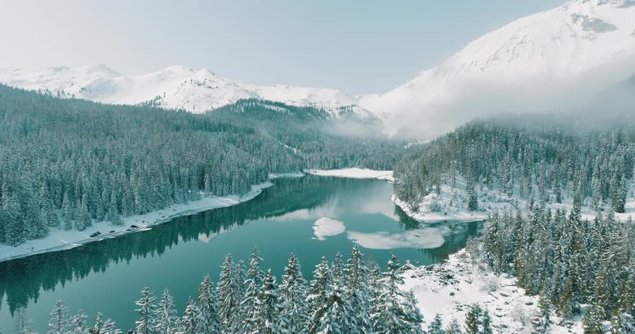 A forest blanketed in winter snow surrounds a lake at its center with a snowcovered mountain range rising in the background under the midday light