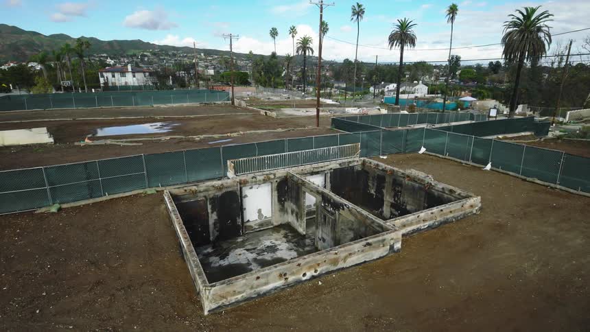 An aerial view shows the concrete foundation and basement ruins of a demolished structure on a dirt lot surrounded by green construction fences under a bright blue sky