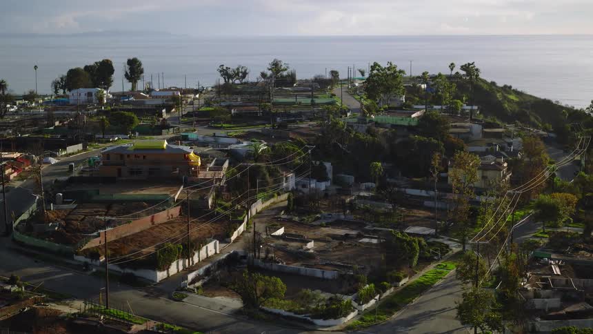 An elevated view looks down over a quiet coastal hillside neighborhood of scattered homes winding roads and power lines interspersed with trees and empty lots all bathed in soft afternoon light as the calm ocean stretches endlessly across the horizon