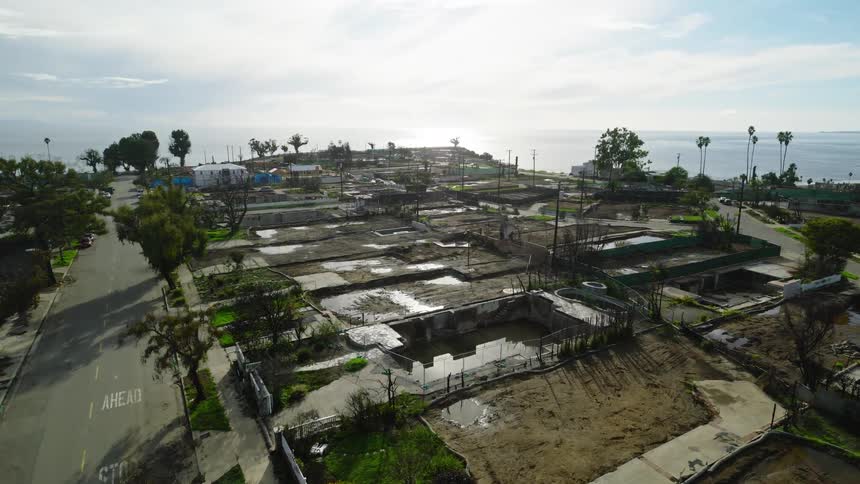 An elevated view surveys a quiet coastal neighborhood with empty foundations fenced lots and sparse trees where sunlit streets lead toward the shimmering ocean on the horizon under a pale open sky