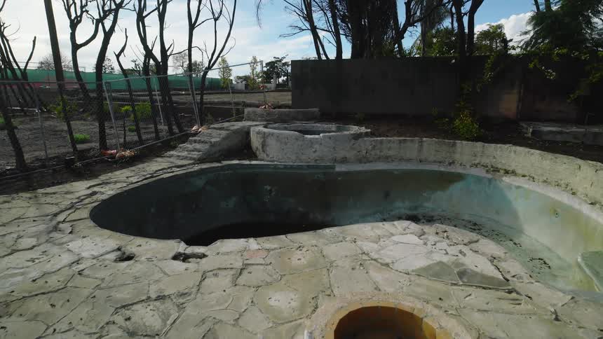 A drained kidneyshaped concrete pool with stained and peeling surfaces is set into cracked stone paving with a small circular fountain basin in the foreground bare trees and fencing around the perimeter and scattered debris visible under daylight