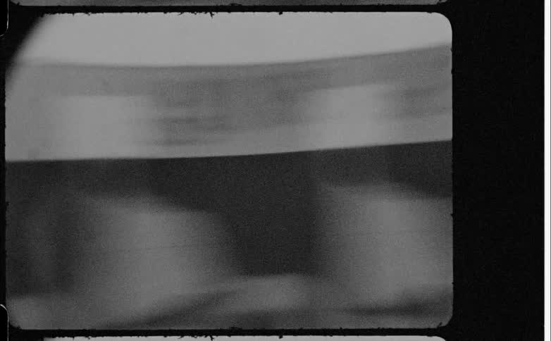 Grainy black and white vintage video captures an abstract blurred foreground view of a film strip with holes visible on the sides