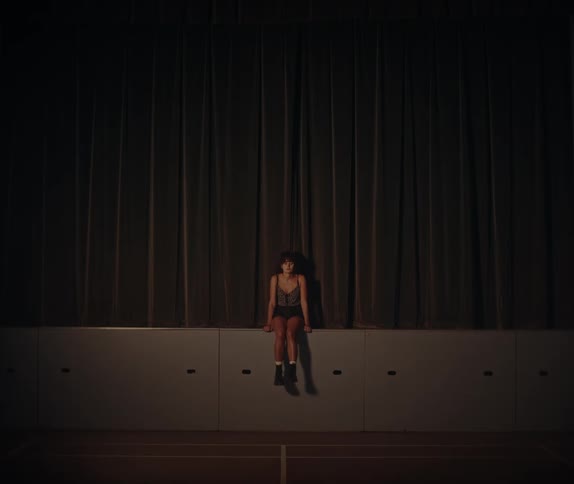 A low angle view shows a woman with dark hair and shorts sitting with her feet dangling on a raised stage area against a dark theater curtain