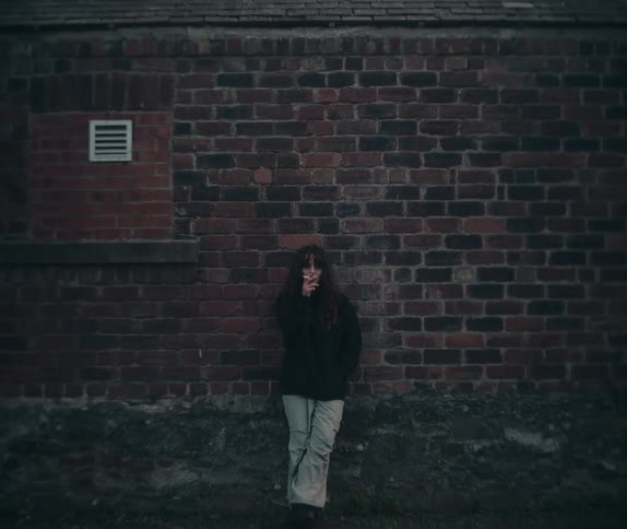 A young woman with long dark hair stands against a dark red brick wall smoking a cigarette on a somber day