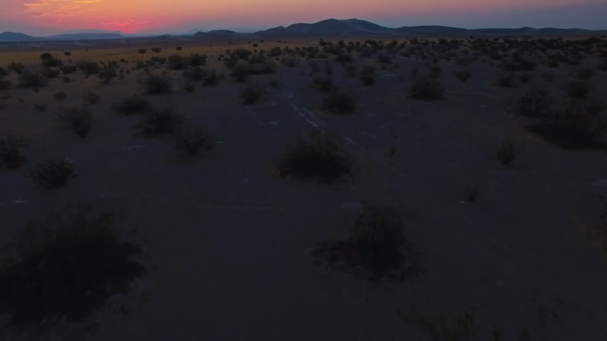 The camera slowly rises from a dark arid landscape dotted with low desert shrubsthe shot reveals a wide vast desert plain at twilight with the warm colors of the setting sun illuminating the distant hills