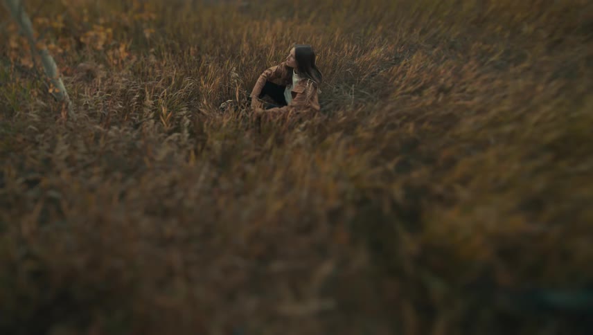 A young woman is seated in a patch of tall dry brown grass looking up and slightly to her leftshe is wearing a light brown jacket and dark pants and the scene has a soft warm autumnal color palette with a shallow depth of field