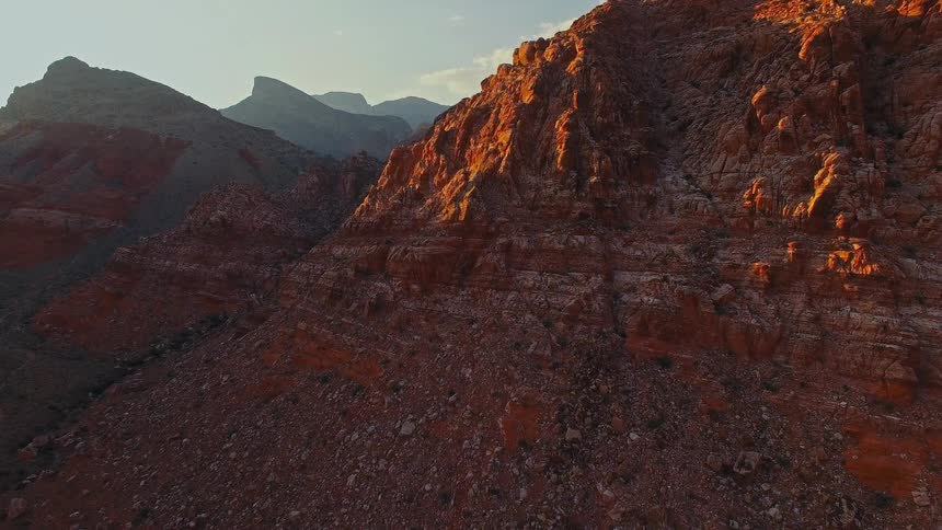 Golden hour sunlight illuminates the rugged peaks of a vast red rock mountain range in a slowmoving aerial shot
