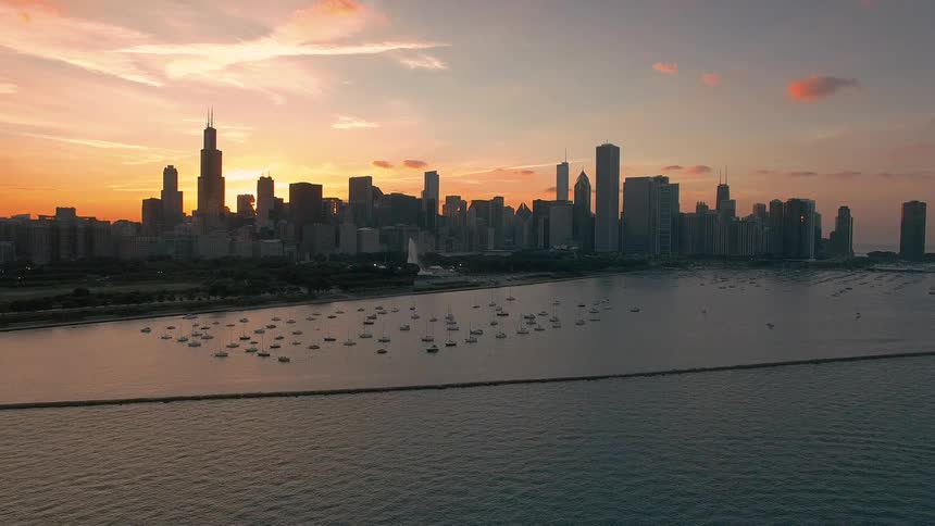 The video is a wide elevated shot of a major city skyline at sunset with a large body of water in the foregroundthe city which includes the recognizable willis tower sears tower is silhouetted against a sky painted with warm orange pink and ye