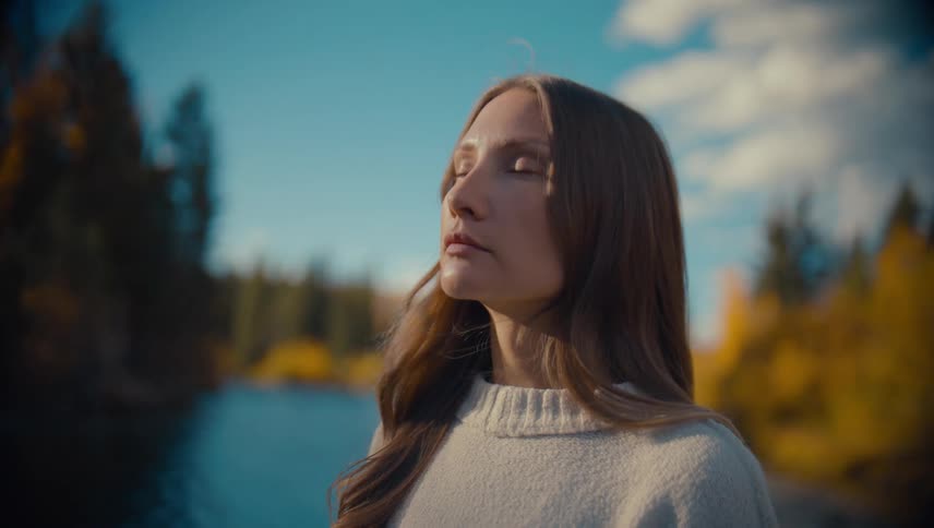 A serene woman closes her eyes while standing near a river amidst colorful autumn foliage