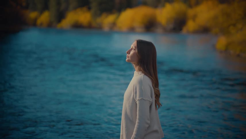 A young woman with long brown hair wearing a lightcolored sweater stands in profile near a body of flowing blue watershe is looking off into the distance with a blurred backdrop of trees showing warm yellow autumn foliage