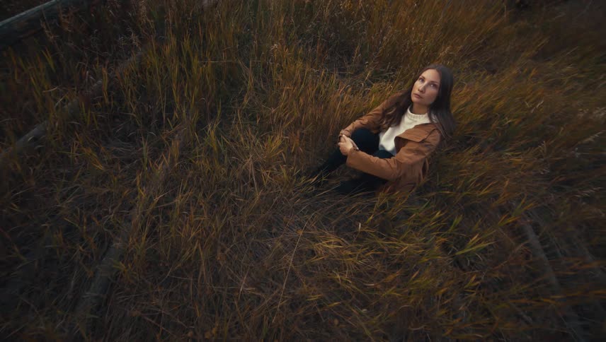 A woman sits in a field of tall dry grass gazing thoughtfully upward