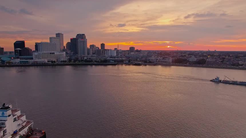 A large cargo ship sails across a river at sunset with a city skyline glowing in the background