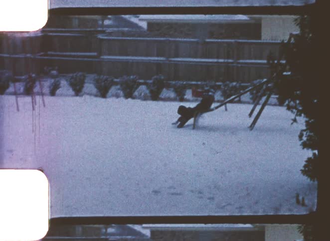 A child slides down a backyard playground slide at midday wearing a winter jacket and boots with the ground covered in snow