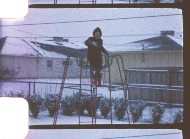 A young boy stands atop a play structure and waves while it snows