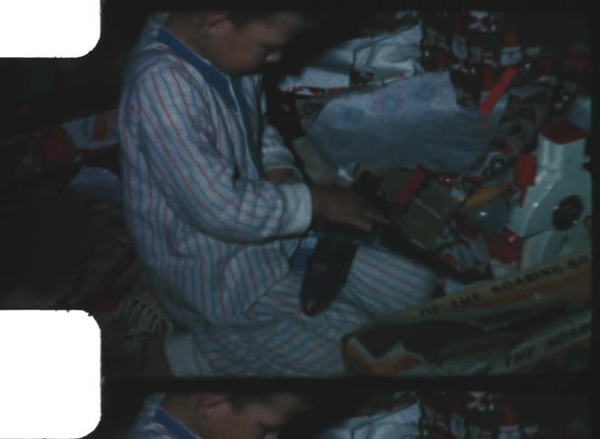 A person in striped pajamas is focused on opening a gift that appears to be packaged in a gray cardboard box the surrounding area is cluttered with wrapping paper and other partially opened gifts indicating a holiday or birthday celebration
