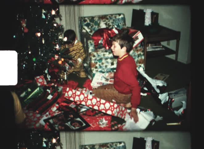 Two young boys open presents on a floor scattered with wrapping paper near a decorated christmas tree