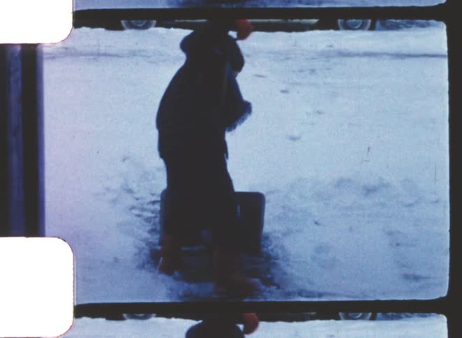 A person wearing dark winter clothes and a hood is shoveling snow on a street or driveway a vintage twotone light and dark station wagon is parked in the snowy background behind them