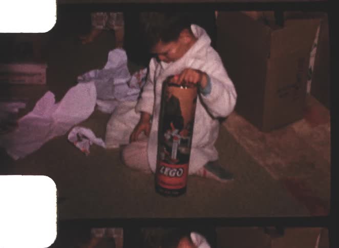 A close view shows a small child in a white robe sitting on the floor opening a tall cylindrical lego tube container with wrapping paper nearby