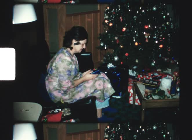 A middle aged woman in a floral print shirt and blue pants sits on a couch opening a gift near a decorated christmas tree