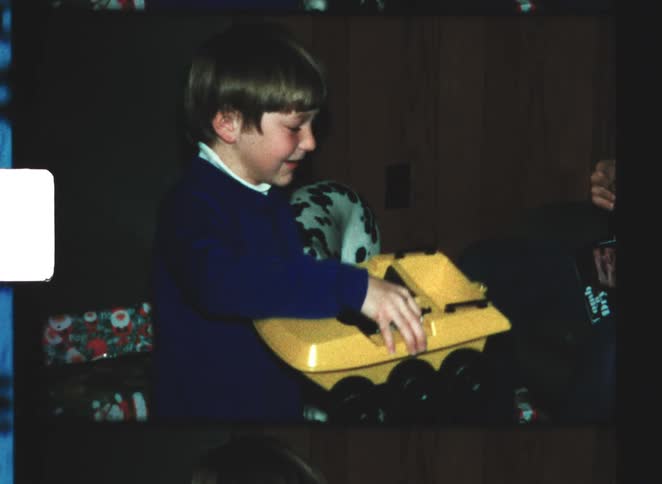 The video is a short clip showing a young boy with light brown hair dressed in a blue sweaterhe is sitting indoors likely opening or examining a gift which appears to be a large yellow toy truck or vehiclethe setting is dark and the boy is conc