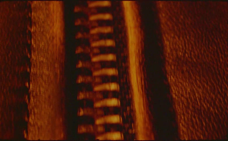 A close view of a shiny metal zipper on brown leather jacket fabric is revealed under warm amber lighting with deep cinematic grain
