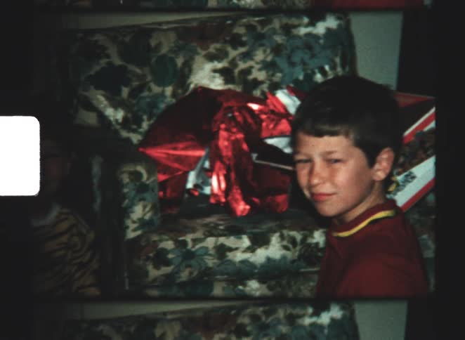 A boy in a red shirt with yellow trim opens presents next to a second boy in a striped shirt near a floral armchair and a decorated christmas tree