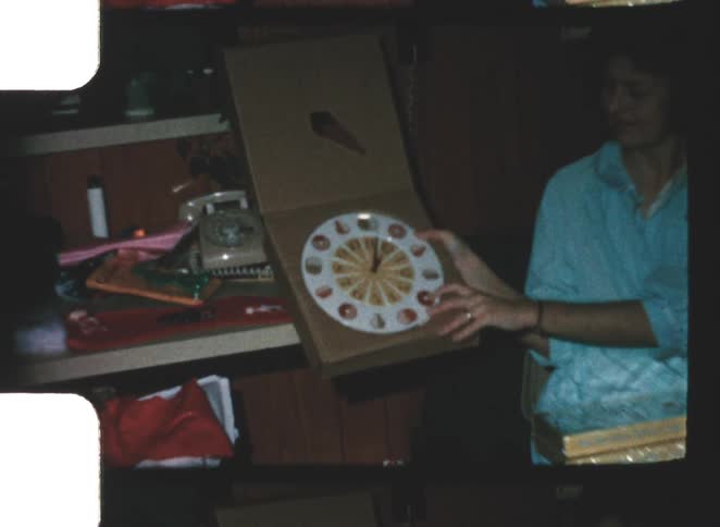 Vintage 1960s color video captures a happy woman in a light blue shirt holding an open box containing a clock in a dimly lit room with wood paneling
