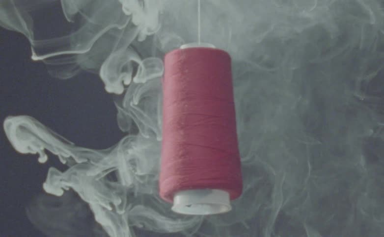 A spool of pink thread hangs in a dark space with swirling white smoke moving around it from below creating a mysterious atmosphere
