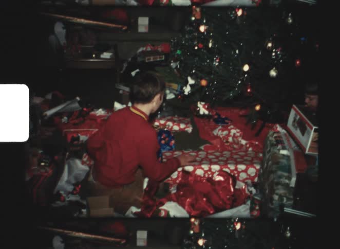 Children open presents in front of decorated christmas tree in room with wrapping paper on the floor